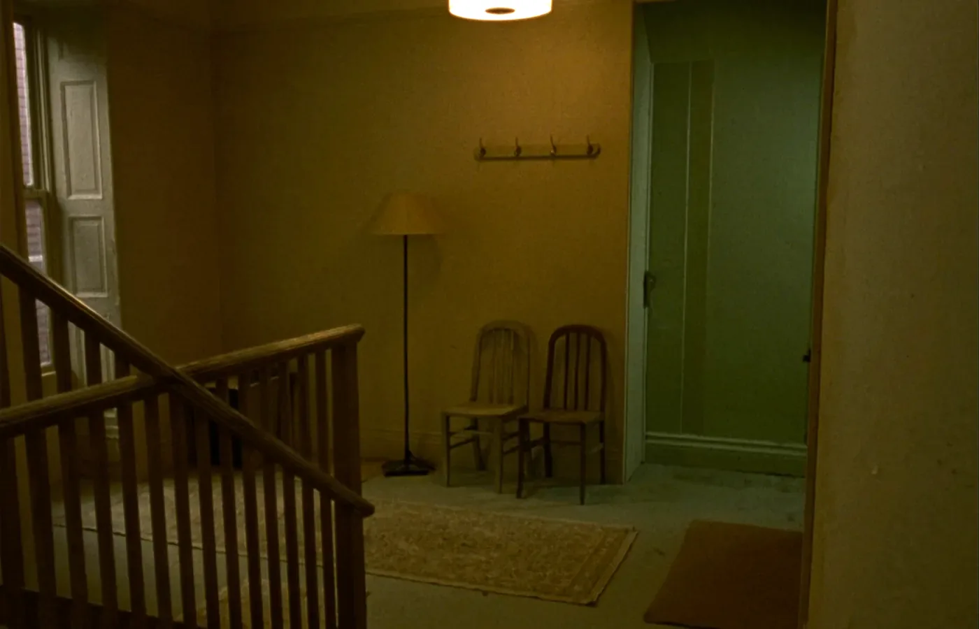 A small waiting area at the top of a wooden staircase, with two plain chairs, a coat rail, a floor lamp and a closed panelled door.